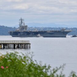 A large aircraft carrier sailing in calm waters with a pier in the foreground and hills in the background.