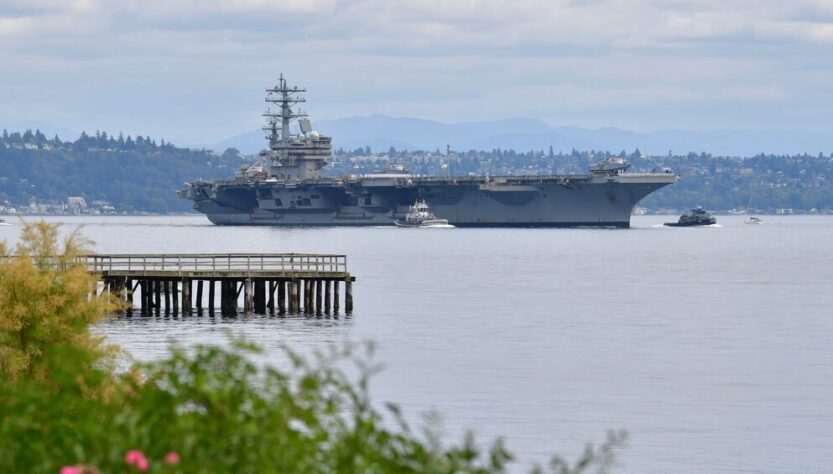 A large aircraft carrier sailing in calm waters with a pier in the foreground and hills in the background.